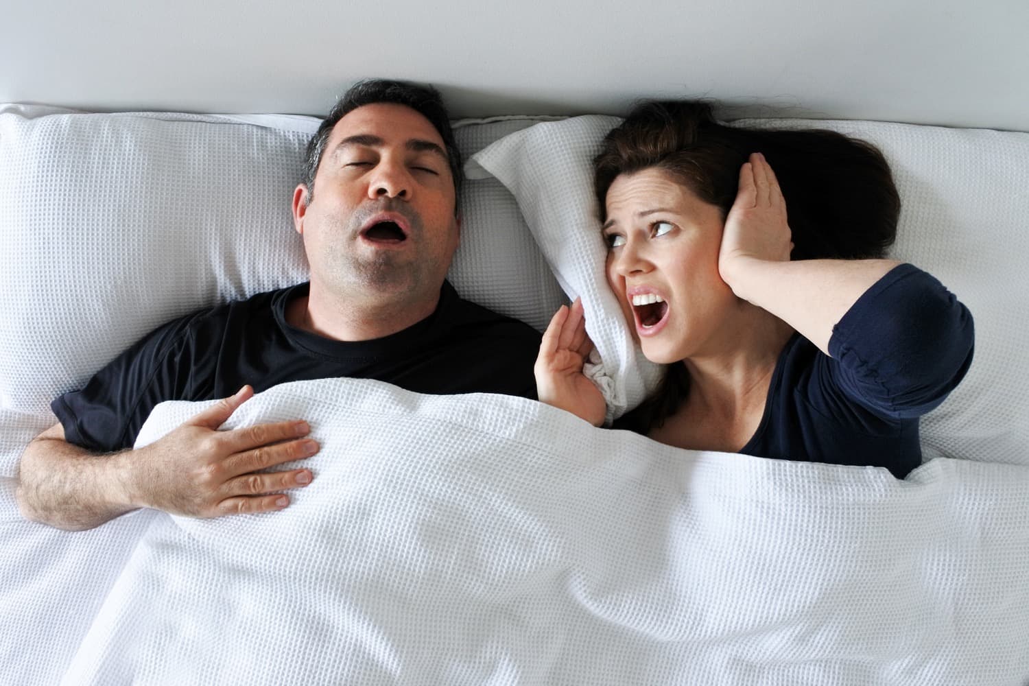 Women in bed with partner covering her ears with a look of frustration over partner snoring loudly.